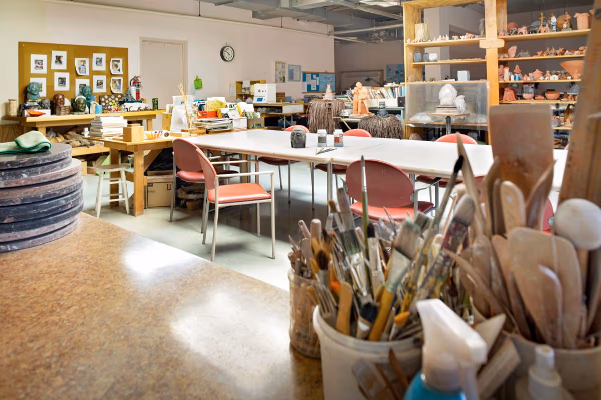 A well-lit art studio with a large table surrounded by red chairs. Various art supplies, including paintbrushes and sculpting tools, are organized in containers in the foreground. Shelves filled with pottery and sculptures line the walls, and there are several busts and art pieces displayed on tables and shelves. The room has a creative and organized atmosphere.