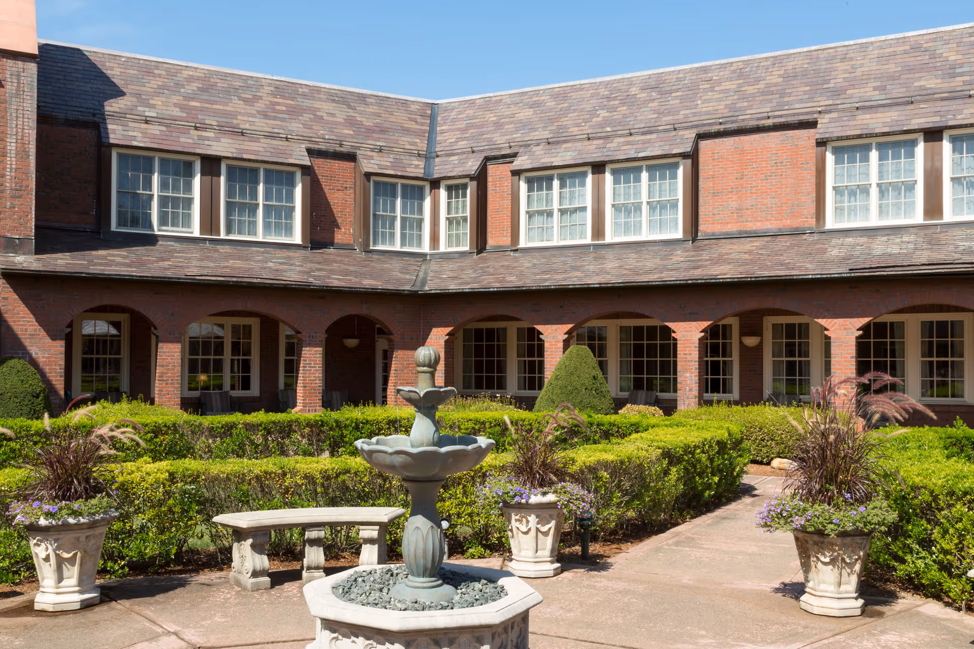 A courtyard garden area with neatly trimmed hedges, a stone fountain in the center, stone benches, and potted plants. The courtyard is surrounded by a two-story brick building with multiple windows and arched walkways.