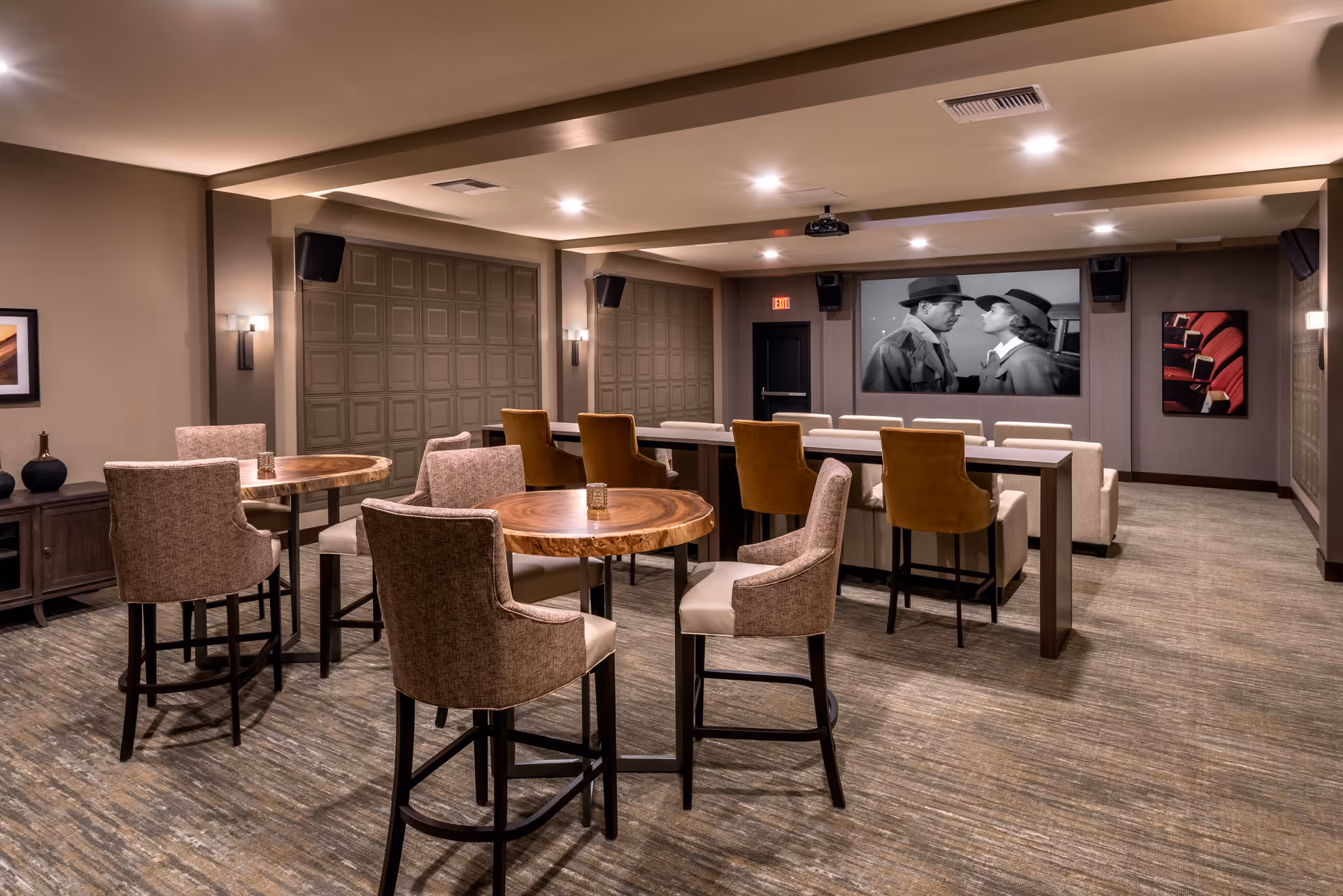 A cozy media room with a large screen showing a black-and-white movie. The room has several high tables with cushioned chairs and a row of bar-height chairs facing the screen. The walls are decorated with framed pictures, and the lighting is warm and recessed in the ceiling.