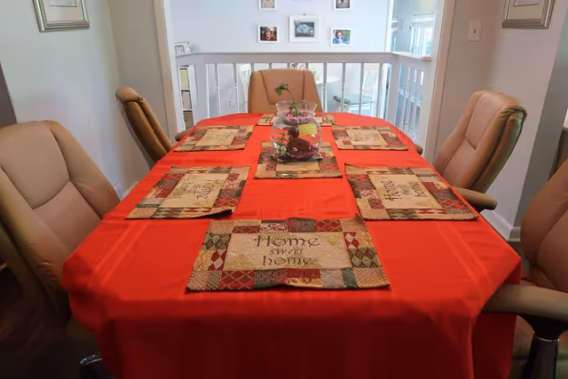 A dining table covered with a red tablecloth and six beige cushioned chairs around it. The table has six placemats with a patchwork design and the words 'Home sweet home' embroidered on them. In the center of the table, there is a glass bowl with decorative items inside. The background shows a white railing and framed pictures on the wall.
