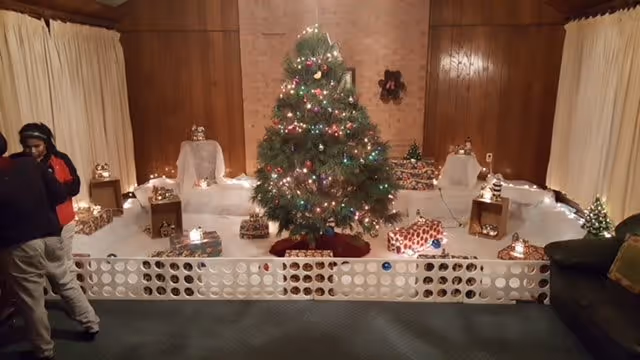 A decorated Christmas tree with colorful lights and ornaments is displayed in the center of a room with wood-paneled walls and beige curtains. The tree is surrounded by wrapped presents and small lit decorations placed on white cloth-covered platforms. A white plastic barrier is set up in front of the display. Two people are partially visible on the left side of the image, and a dark green couch is on the right side.