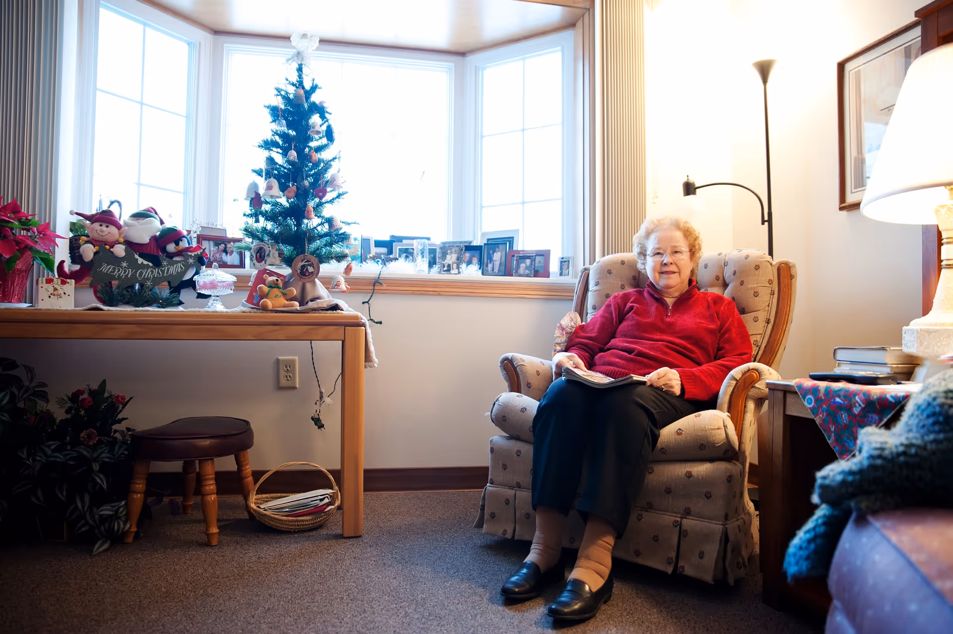 An elderly woman wearing a red sweater sits comfortably in a cushioned armchair in a cozy living room. To her left, there is a wooden table decorated with Christmas ornaments, including a small Christmas tree and festive figurines. The room is softly lit by a floor lamp and a table lamp, with a large window in the background letting in natural light. Various framed photos are displayed on the windowsill.