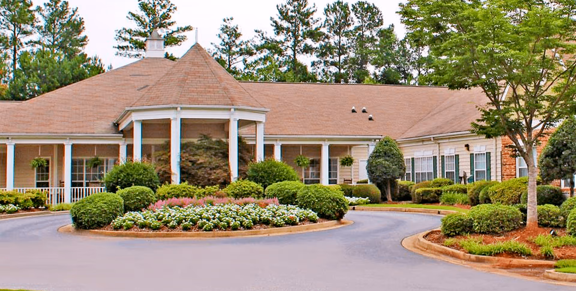 Exterior view of a single-story building with a tan roof and white columns, surrounded by well-maintained bushes, trees, and flower beds, with a circular driveway in front.