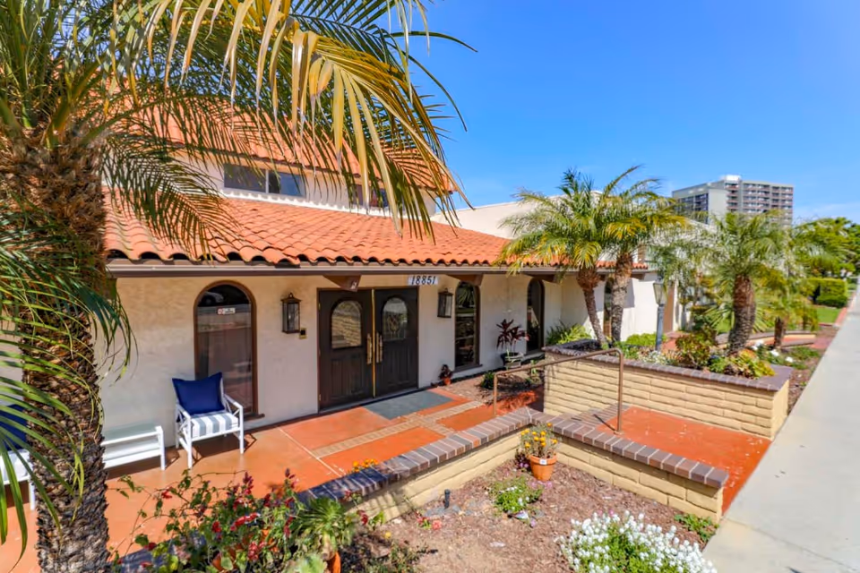 Front entrance of a Mediterranean-style assisted living building with double doors, a terracotta tile roof, palm trees, and a paved walkway.
