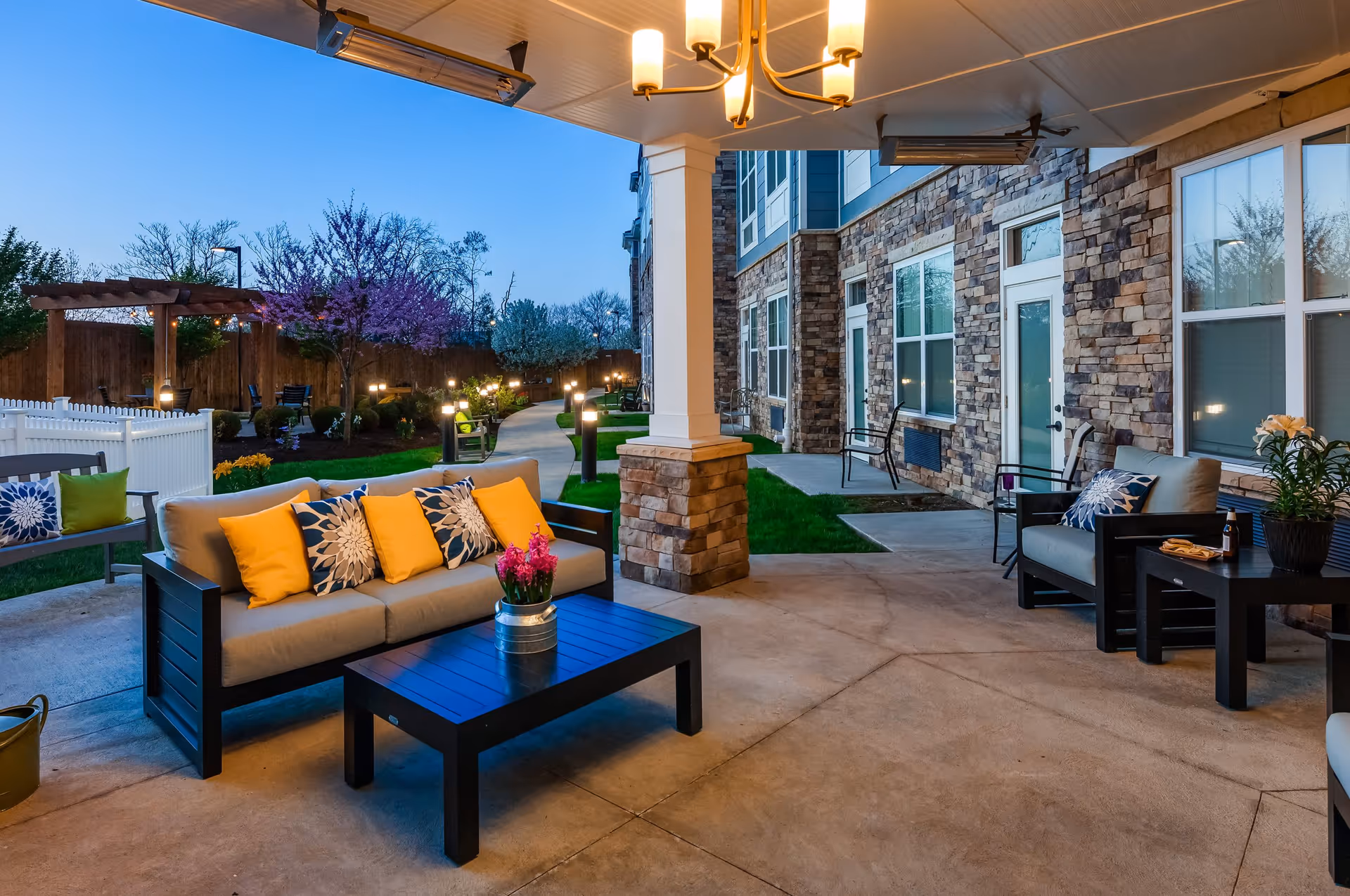 Covered outdoor patio area with cushioned seating including a sofa and armchairs, decorated with yellow and blue patterned pillows. A black coffee table with a potted pink flower sits in front of the sofa. The patio overlooks a landscaped garden with a winding pathway, small trees, and outdoor lighting. The building exterior features stone walls and multiple windows and doors.