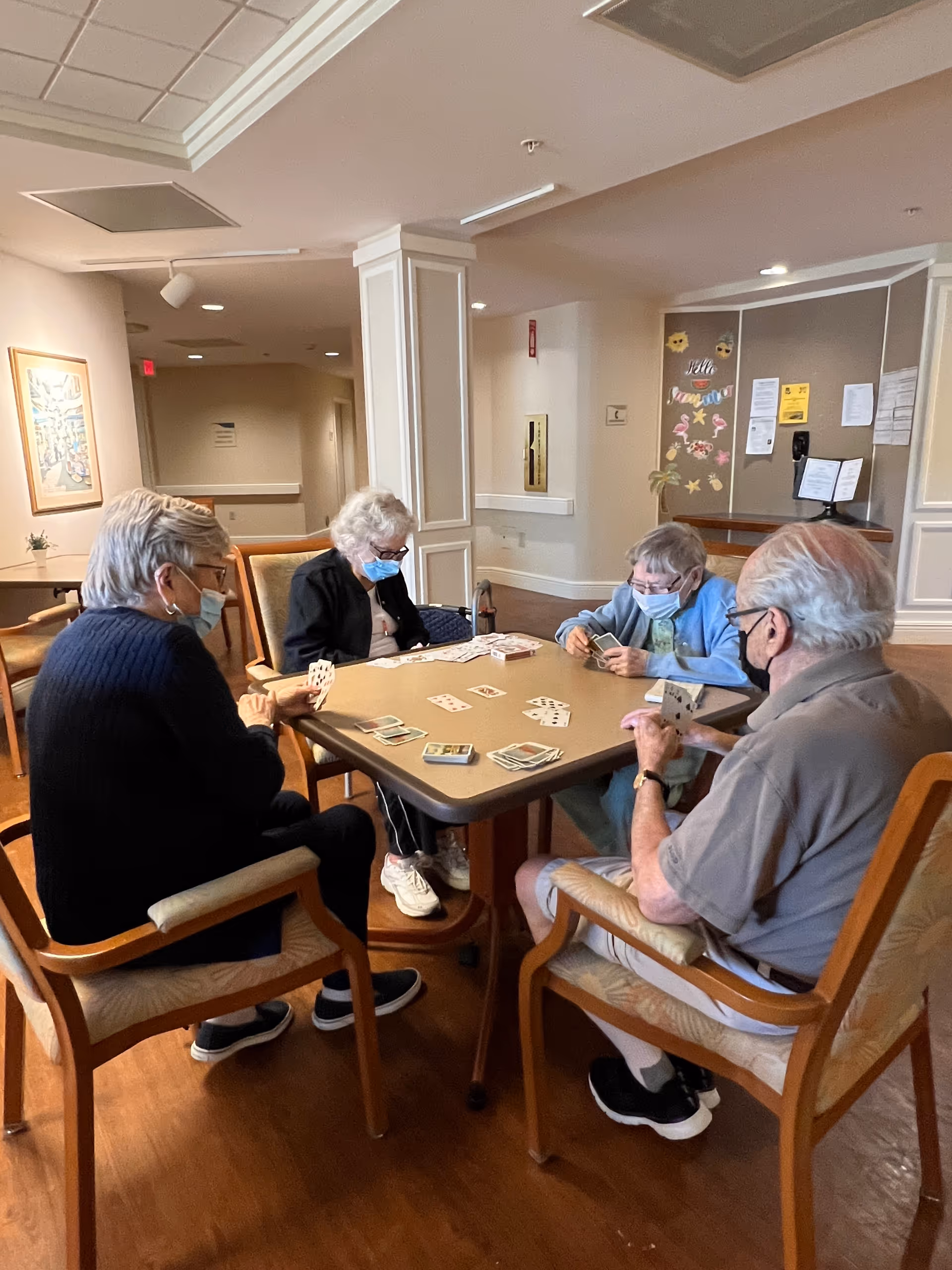 Four elderly individuals wearing face masks sitting around a square table playing a card game in a well-lit room with wooden floors and beige walls. The room has comfortable chairs, a bulletin board with decorations and notices, and a framed picture on the wall.