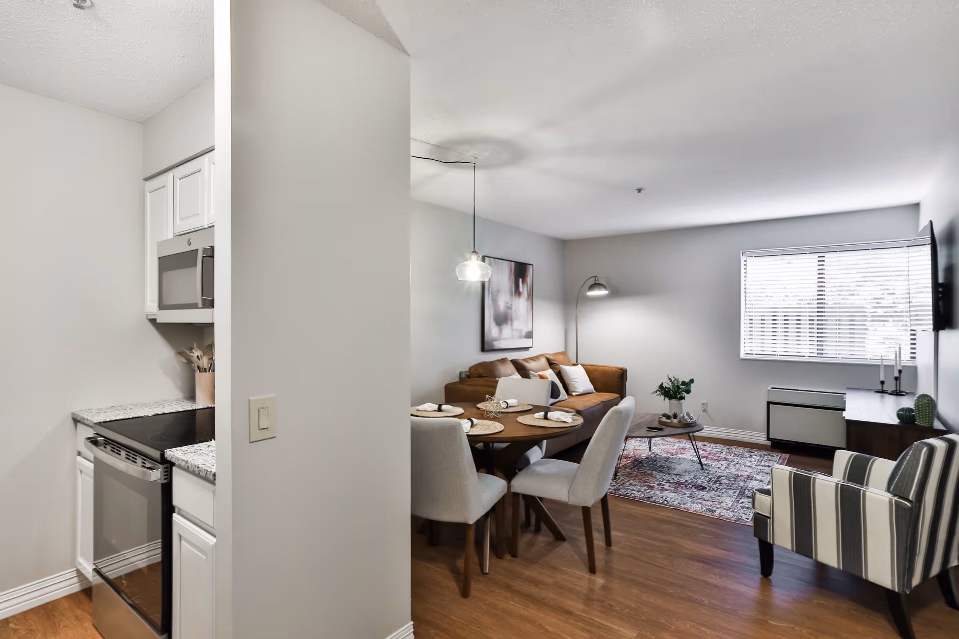 Interior view of an apartment showing a small kitchen area with a stove and microwave on the left, and an open living and dining area on the right. The dining area has a round wooden table set with four chairs and place settings. The living area features a brown leather sofa, a striped armchair, a coffee table with plants, a floor lamp, a wall-mounted TV, and a window with blinds letting in natural light.