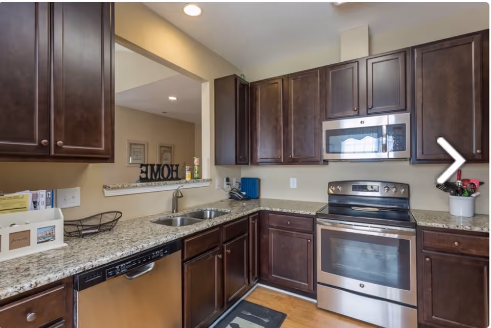Modern kitchen with dark wooden cabinets, granite countertops, stainless steel appliances including a dishwasher, oven, and microwave. There is a double sink under a pass-through window with a decorative HOME sign on the ledge. The floor is wooden and the kitchen is well-lit with recessed lighting.