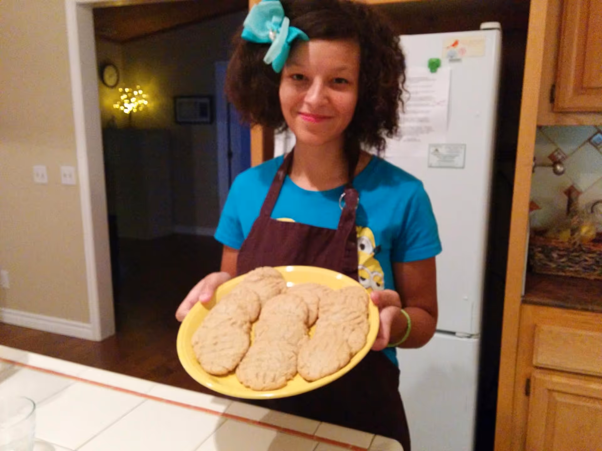A young woman with curly hair and a large blue bow is standing in a kitchen holding a yellow plate with six large cookies. She is wearing a blue shirt and a dark apron. Behind her is a white refrigerator and wooden kitchen cabinets.