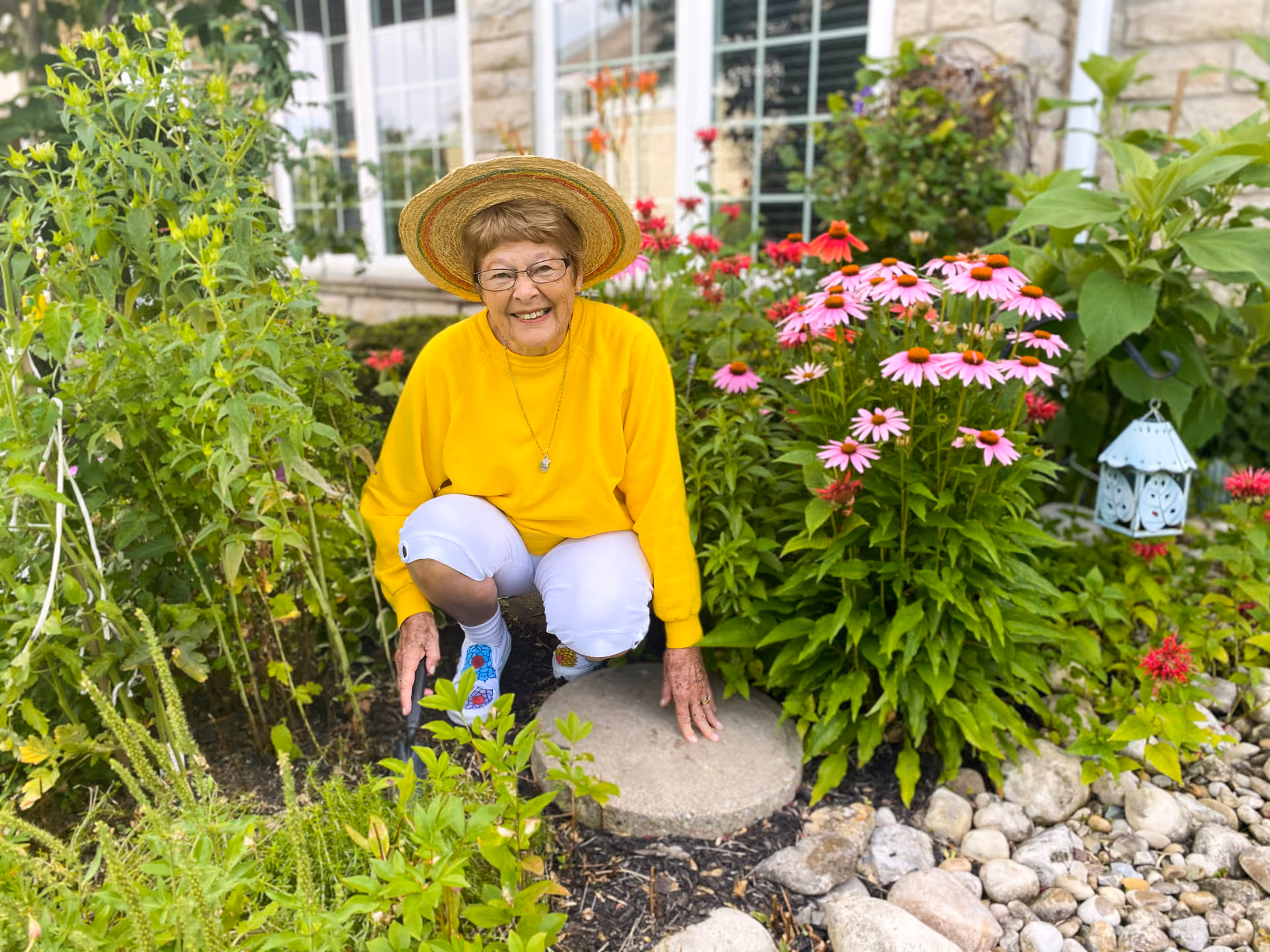 An elderly woman wearing a yellow sweater, white pants, and a straw hat is crouching in a garden surrounded by green plants and pink flowers. She is smiling and appears to be enjoying the outdoor setting near a building with large windows.