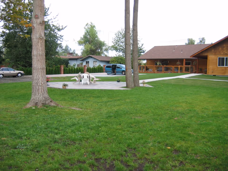 Outdoor area with green grass, two large trees, a wooden deck with white plastic chairs and a table, a wooden building with a porch, and a driveway with parked cars in the background.