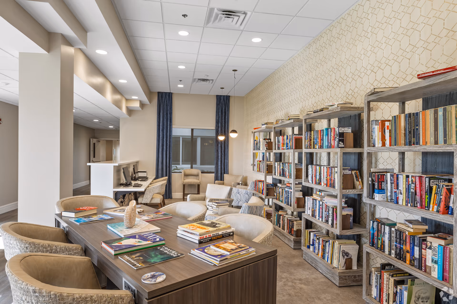 A well-lit library or reading room with multiple bookshelves filled with books along the right wall. In the center, there is a large wooden table with several chairs around it and books spread out on the table. In the background, there are two armchairs near a window with dark curtains, and a small desk with two computer stations on the left side.