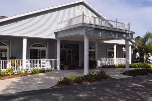 Front entrance of Arcadia Oaks Assisted Living, a light-blue building with a covered porte-cochère, white columns, and a wraparound porch and railings.