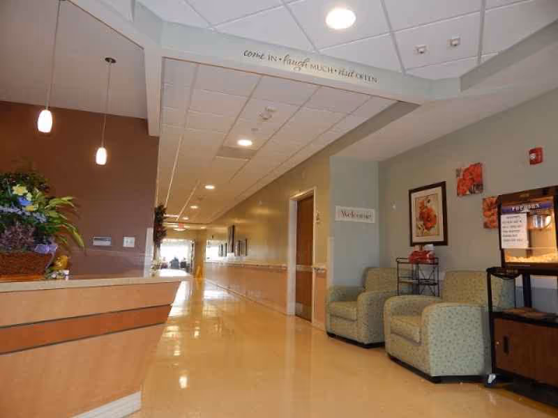 Interior hallway of a senior care facility with a reception desk on the left, two armchairs and framed floral artwork on the right wall, and a popcorn machine. The hallway is well-lit with ceiling lights and pendant lights above the reception desk. A welcoming sign is visible on the wall near the chairs.
