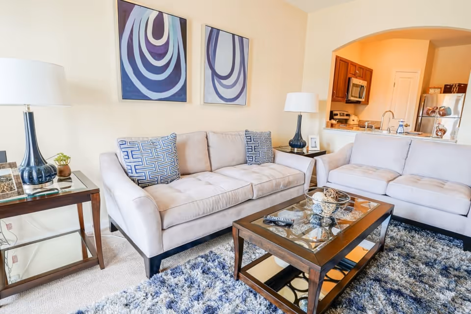 A bright living room with two beige sofas, each with blue patterned pillows, a glass-top wooden coffee table with decorative items, a blue and white shag rug, two matching table lamps on wooden side tables, and two abstract blue and purple paintings on the wall. The room opens into a kitchen area with wooden cabinets and stainless steel appliances.