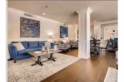 A spacious living room area with a blue sofa, patterned armchairs, a wooden coffee table on a light-colored rug, and abstract paintings on the walls. The room has hardwood flooring and white walls with recessed lighting. In the background, there is a dining area with a table and chairs.