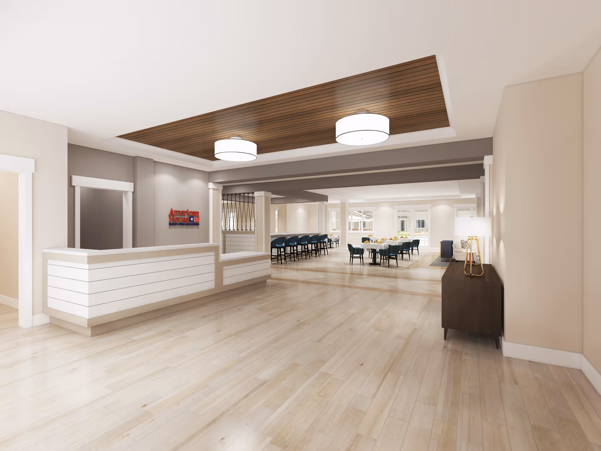 Interior view of a senior living facility lobby area with a white reception desk on the left, a wooden ceiling panel with two round ceiling lights, and a dining area with tables and chairs in the background. There is a small dark wooden cabinet with a lamp on the right wall.