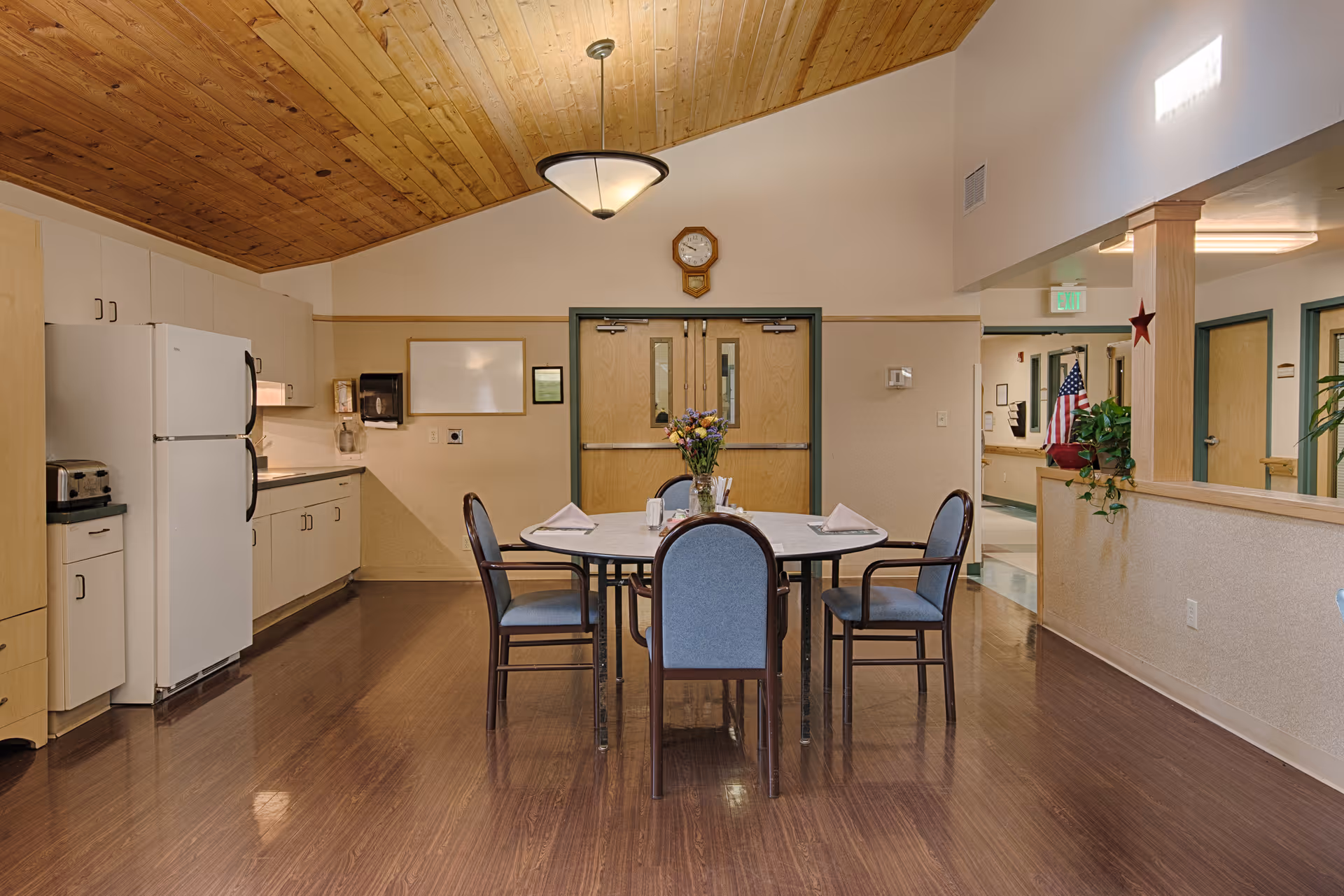 Interior view of a dining area in a senior living facility with a round table set for four, a flower vase centerpiece, wooden ceiling, and a kitchen area with white cabinets and appliances in the background.