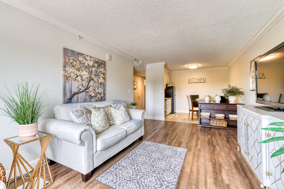 A bright and cozy living room area with a light gray sofa adorned with patterned cushions, a small side table with a potted plant, a decorative rug on wooden flooring, and a large floral painting on the wall. In the background, there is a dining area with a wooden table and chairs, a dark wooden sideboard with decorative items and baskets, and a kitchen area with white cabinets and appliances. The word 'KITCHEN' is displayed on the wall above the dining area.