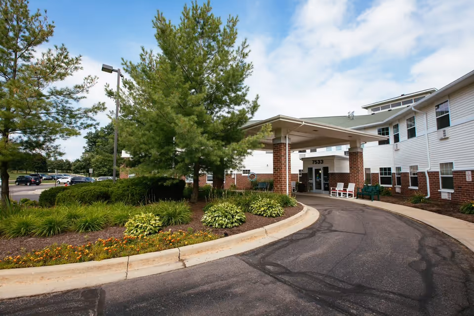Entrance to Woodland Village senior living facility with a covered drop-off area supported by brick columns. The building is white with brick accents and multiple windows. There are green bushes, trees, and flowers along the driveway, and a parking lot with cars is visible in the background under a partly cloudy sky.