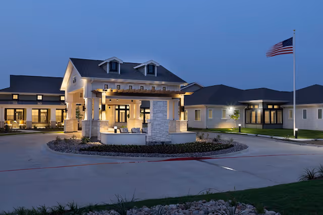 Front entrance of a lit senior living facility at dusk with a covered porte-cochère and an American flag.
