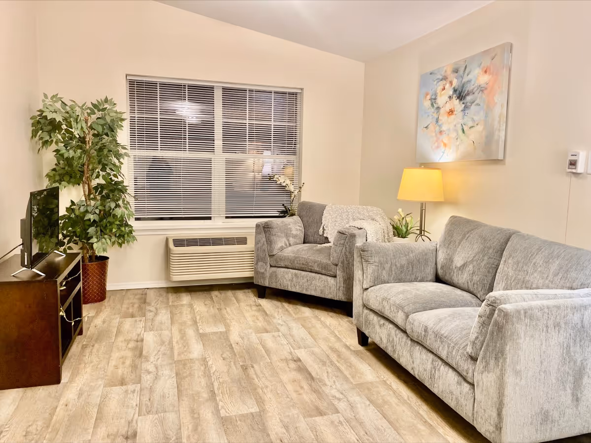 A cozy living room with light wood flooring, a gray sofa and matching armchair, a floor lamp with a yellow shade, a potted plant, a TV on a dark wooden stand, and a large window with white blinds. A floral painting hangs on the wall above the sofa.
