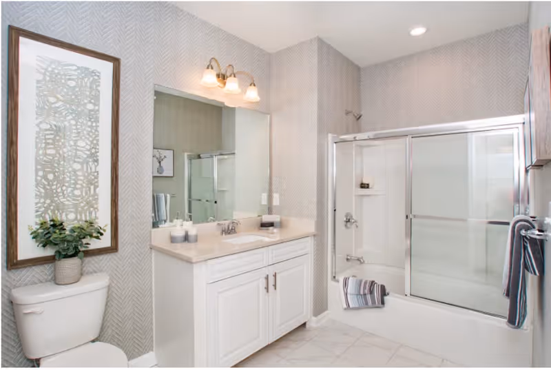 A modern bathroom featuring a white toilet, a vanity with a sink and countertop, a large mirror above the sink, and a bathtub with sliding glass doors. The walls have a subtle patterned wallpaper, and there are decorative elements including a framed artwork above the toilet and neatly folded towels on the bathtub and towel rack.