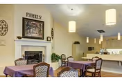 A cozy dining area with several tables covered in purple tablecloths and chairs with patterned upholstery. There is a white fireplace with a framed picture above it and the word FAMILY displayed on the wall. The room is softly lit with hanging pendant lights, and there is a kitchen area visible in the background.