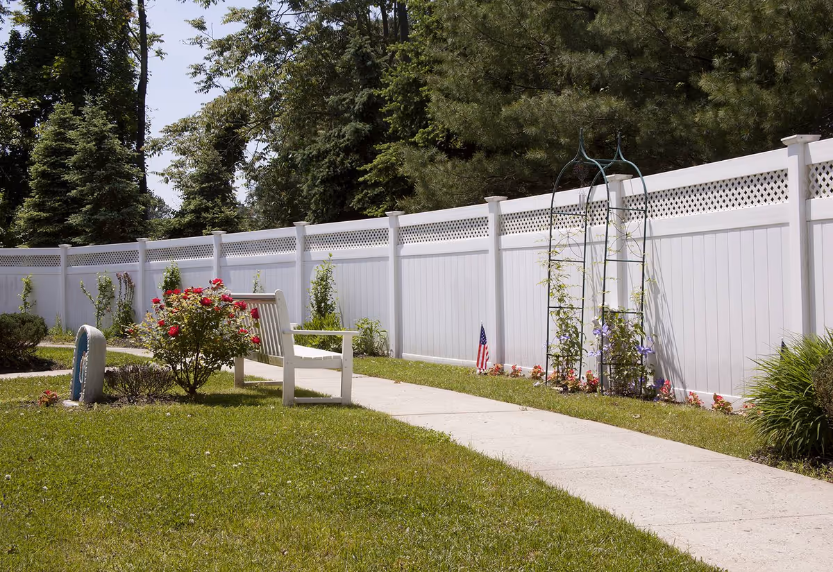 Sunlit courtyard with a white bench along a winding path, flowering shrubs and a tall white privacy fence backed by trees.