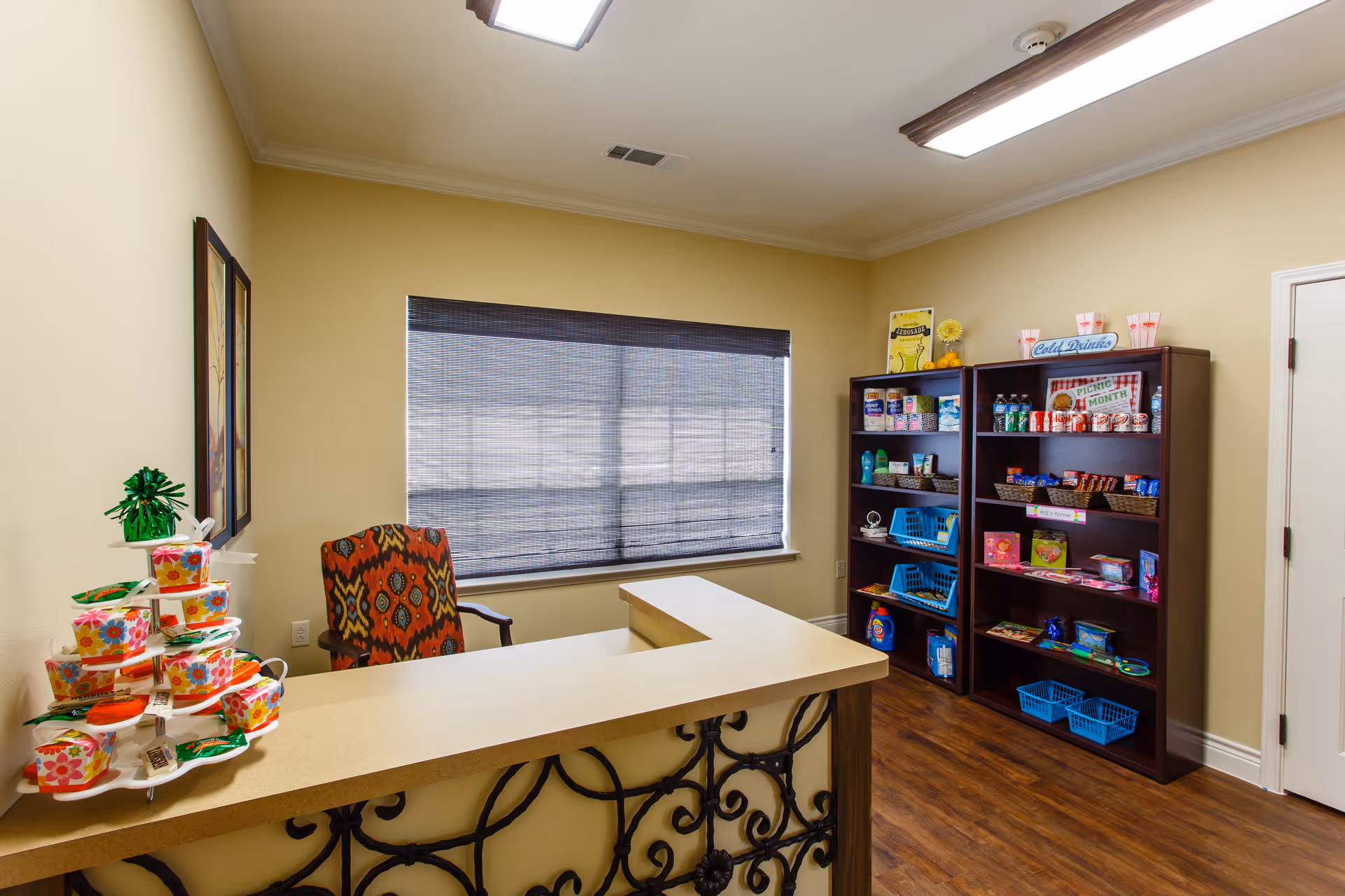 Interior view of a small reception or snack area in an assisted living facility. There is a counter with decorative ironwork on the front, a colorful patterned chair behind it, and two dark wooden shelves stocked with snacks, drinks, and other small items. A window with a closed blind is on the back wall, and the room has wood flooring and beige walls.