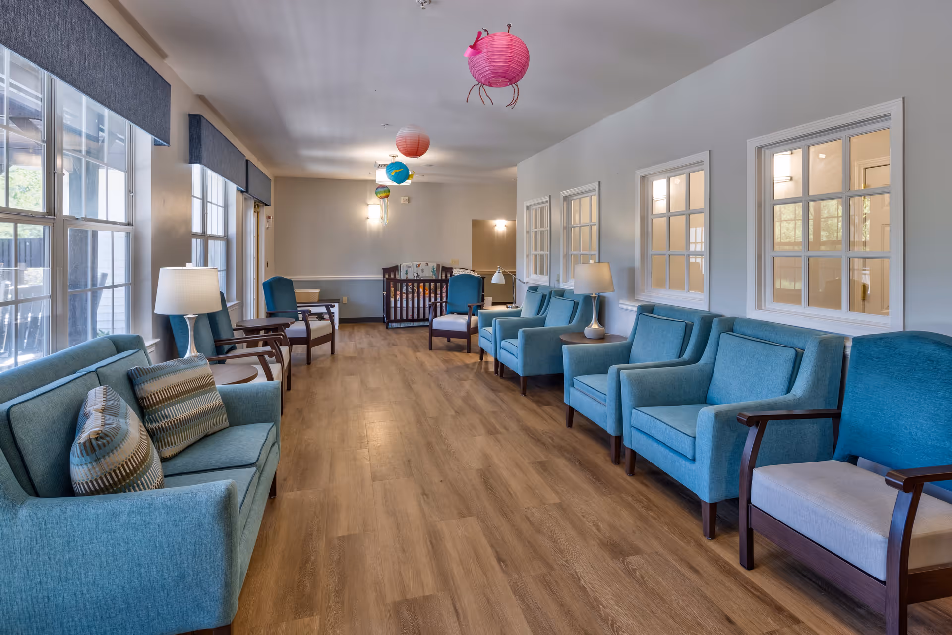 A bright and spacious assisted living facility common area with multiple blue upholstered armchairs and sofas arranged along the walls. The room has large windows on one side letting in natural light, wooden flooring, and colorful paper lanterns hanging from the ceiling. There are lamps on side tables placed between some chairs, and a baby crib is visible at the far end of the room.