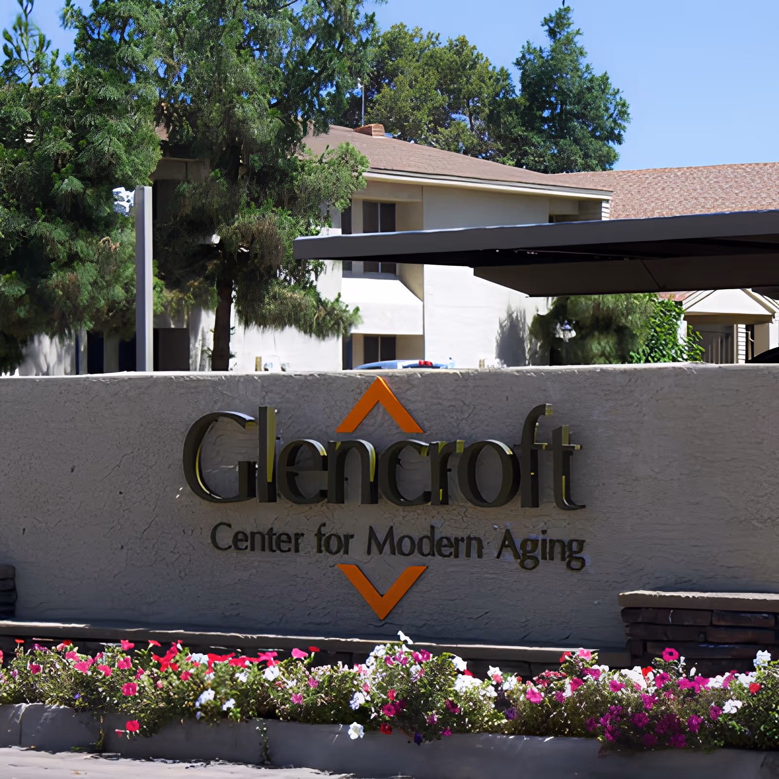 Outdoor view of the entrance sign for Glencroft Center for Modern Aging, with a beige wall, colorful flowers in front, trees, and a building in the background under a clear blue sky.