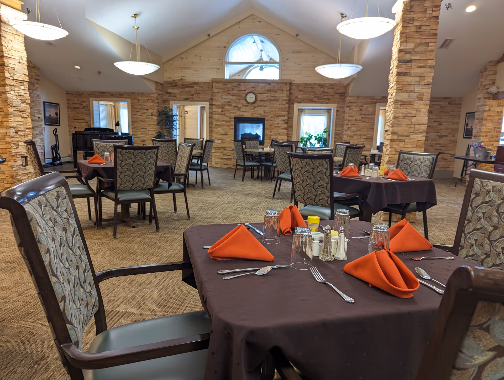 Dining room with multiple tables covered in dark brown tablecloths, each set with orange folded napkins, glasses, and silverware. The room features stone pillars and walls, carpeted floor, large windows, and hanging ceiling lights. A fireplace and a piano are visible in the background.