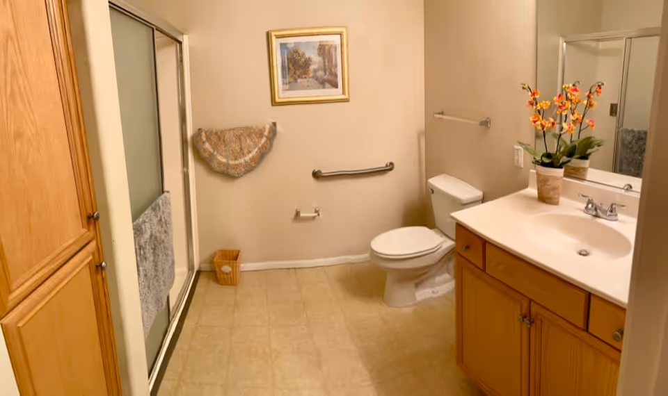 A clean bathroom with a toilet, a sink with a wooden cabinet, a large mirror, a shower with a sliding glass door, a towel rack with a decorative towel, a small wastebasket, and a framed picture on the wall.
