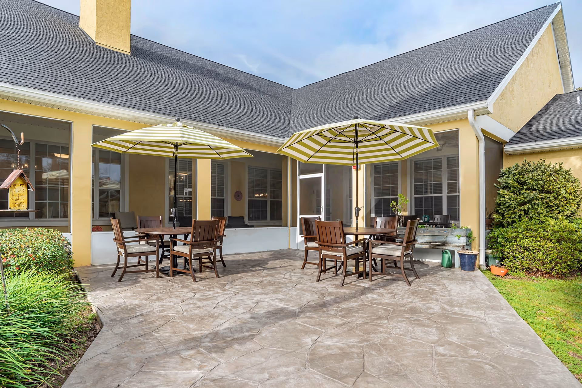 A sunny patio with striped umbrellas, round tables, and chairs outside a beige senior living building.