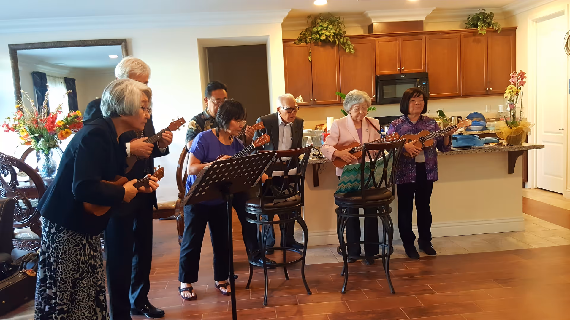 A group of seven elderly people standing in a living room area, playing ukuleles together. The room has wooden floors, a kitchen counter with bar stools, and decorative plants on top of the cabinets. There is a large mirror and a vase with flowers in the background.