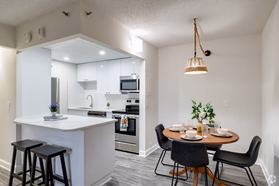 A modern kitchen and dining area featuring white cabinetry, stainless steel appliances including a refrigerator, stove, and microwave. The kitchen has a white countertop island with two black stools. Adjacent to the kitchen is a round wooden dining table set with four black chairs, tableware, and a green plant centerpiece. A woven pendant light hangs above the dining table.