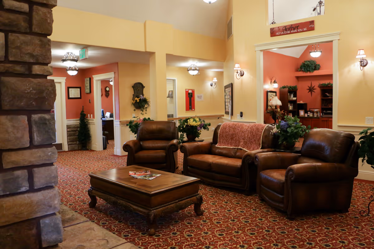 A cozy communal living room with leather sofas and armchairs arranged around a wooden coffee table in a warmly decorated senior facility lounge.