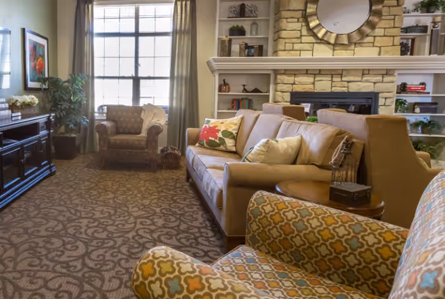 A cozy living room with patterned carpet, a beige sofa adorned with colorful pillows, two patterned armchairs, a stone fireplace with a round mirror above it, built-in shelves with decorative items, a large window with curtains, and a dark wooden cabinet with a plant and framed artwork on the wall.