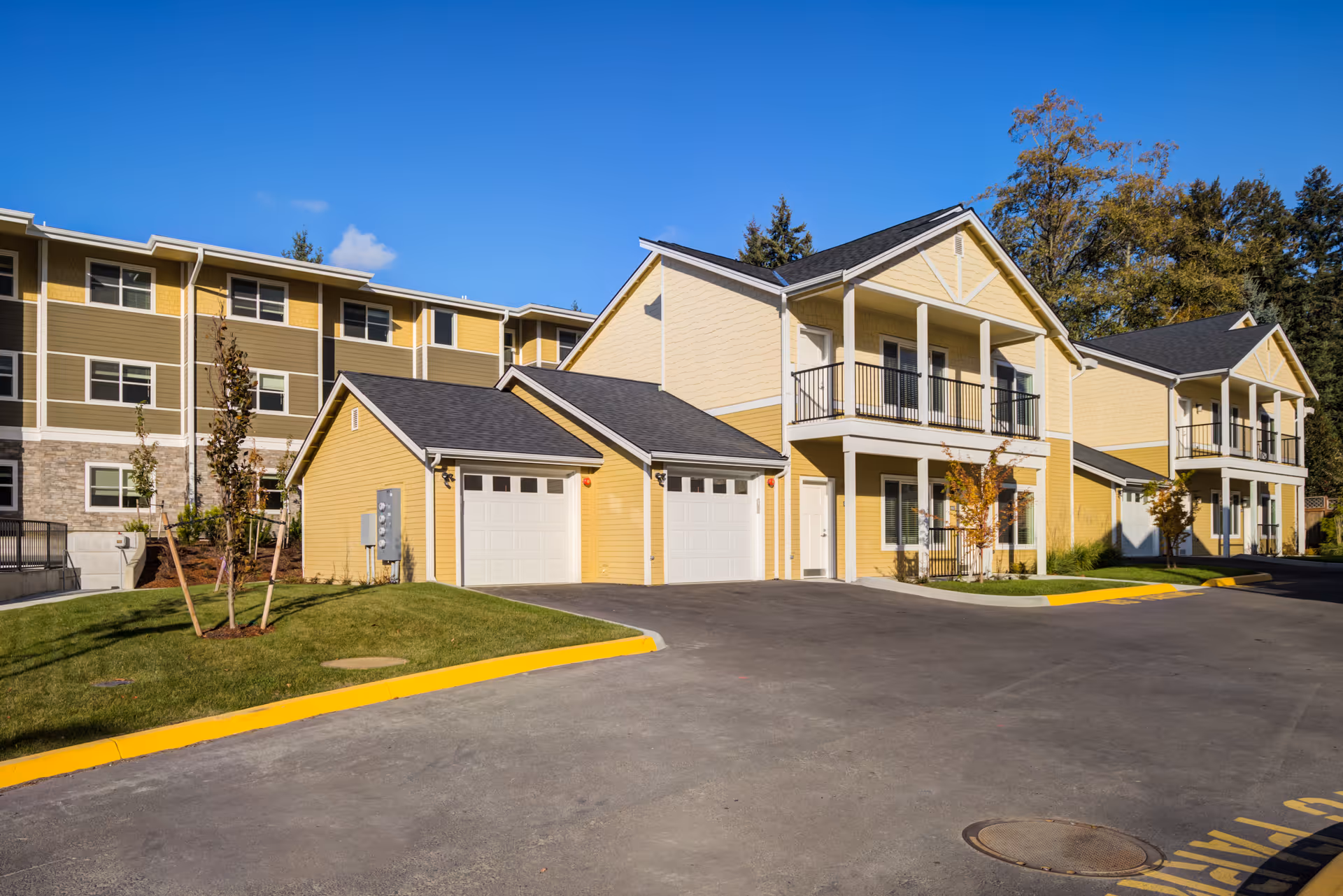 Exterior view of a senior living community featuring yellow two-story buildings with balconies and attached garages, surrounded by a paved driveway and green landscaping under a clear blue sky.