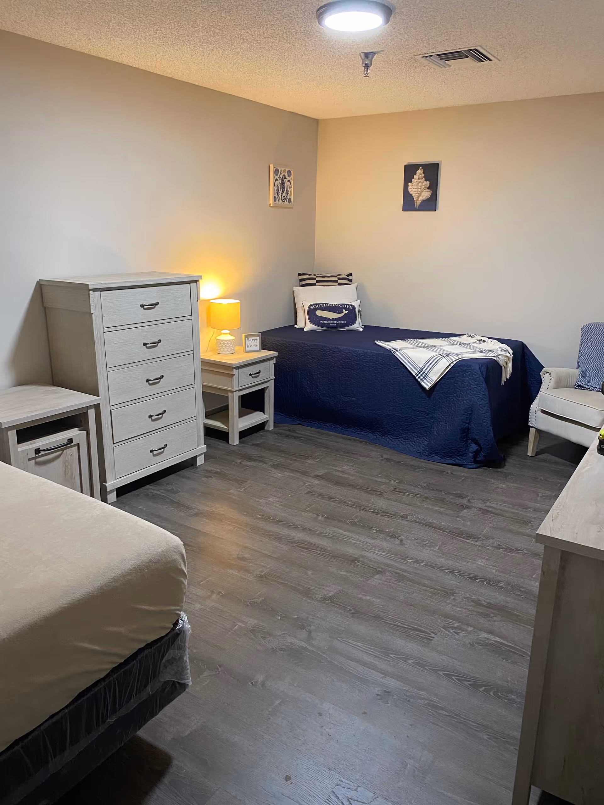A cozy senior living bedroom with two beds, one covered with a navy blue quilt and decorative pillows, and the other partially visible with a beige cover. The room features light-colored wooden furniture including a tall dresser, a nightstand with a lit lamp, and a small cabinet. The walls are painted in neutral tones with two framed artworks hanging. The floor is a dark wood laminate.