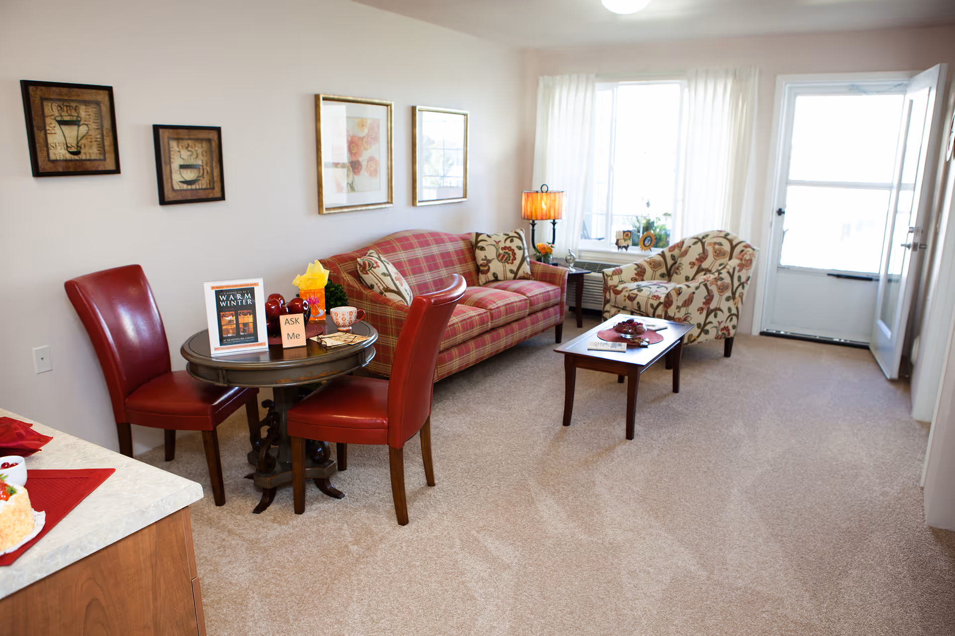 A cozy living room with a plaid red sofa, a floral armchair, a wooden coffee table with decorative items, and a small round table with two red leather chairs. The room has beige carpet, light-colored walls with framed artwork, a window with white curtains, and a door leading outside.