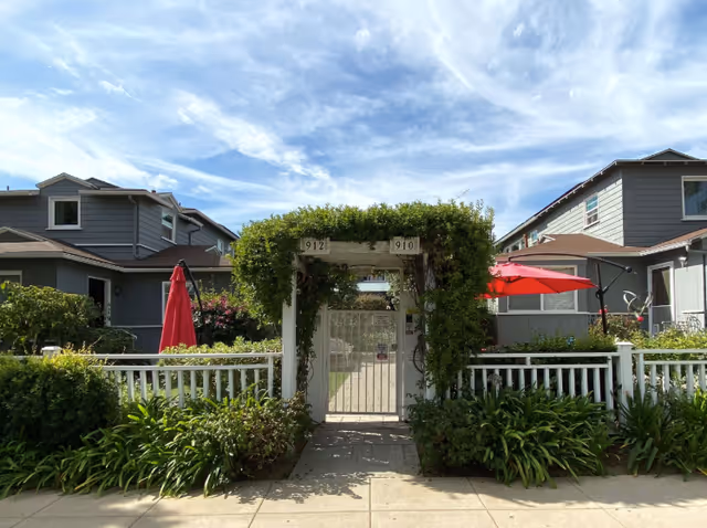 Entrance gate covered with green vines and foliage, leading into a garden area between two gray residential buildings with white fences and red umbrellas in the outdoor seating areas under a partly cloudy sky.