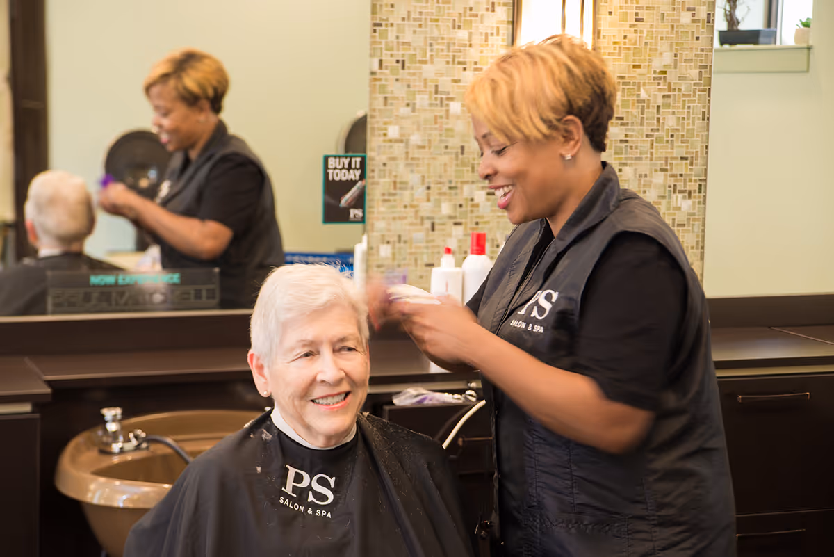 A hairstylist is smiling while cutting the hair of an elderly woman seated in a salon chair. The elderly woman is also smiling and wearing a black salon cape with the letters 'PS' and 'Salon & Spa' printed on it. The background shows a mirror, a sink, and various hair care products.