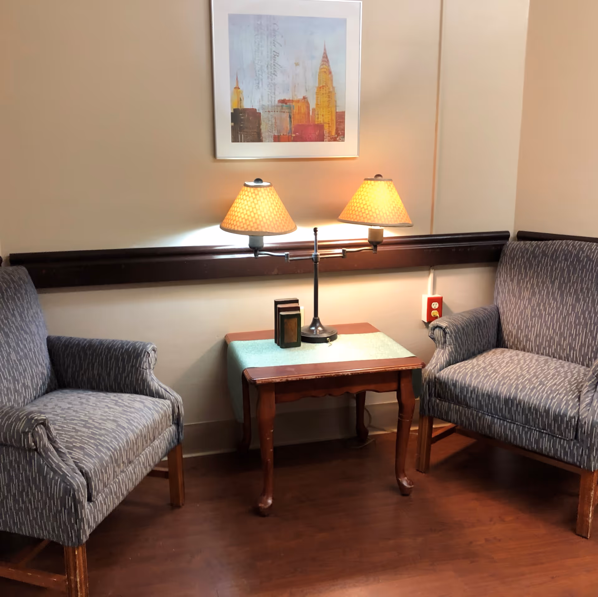 A cozy sitting area with two patterned armchairs facing each other, separated by a wooden side table with a green runner. On the table is a double-shaded lamp and two small decorative books. A framed artwork depicting a cityscape hangs on the beige wall above the table.