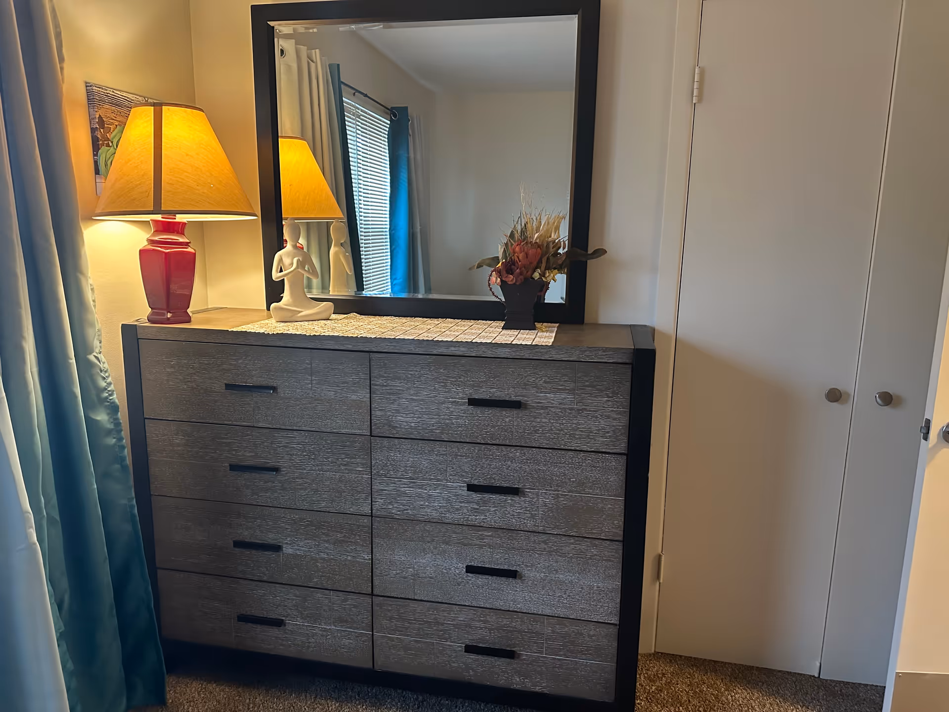 A bedroom dresser with eight drawers, topped with a large rectangular mirror. On the dresser, there is a red lamp with a beige lampshade, a white figurine in a meditative pose, and a small vase with dried flowers. To the right of the dresser is a closed white closet door, and to the left are blue curtains partially covering a window.