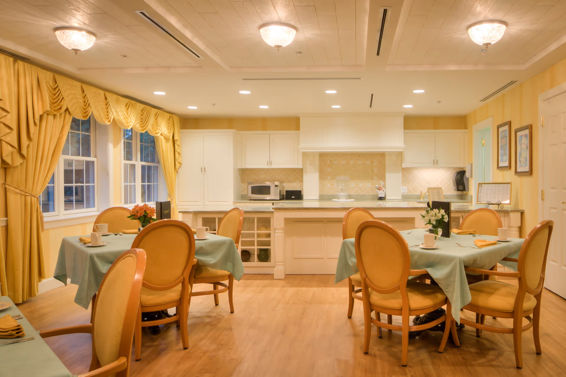 A cozy dining area with two tables covered in light green tablecloths, each surrounded by four wooden chairs with cushioned seats. The room features warm yellow walls, large windows with yellow drapes, and a kitchen area in the background with white cabinets, a microwave, and a countertop. The floor is wooden, and the ceiling has recessed lighting and decorative light fixtures.