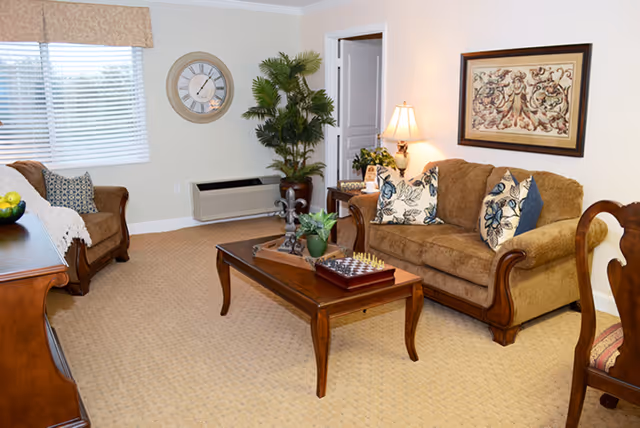 A cozy living room with beige carpet and cream walls featuring a brown upholstered sofa with floral cushions, a matching armchair, a wooden coffee table with a chess set and decorative plants, a side table with a lamp, a large wall clock, a framed artwork, and a potted plant near an open doorway.