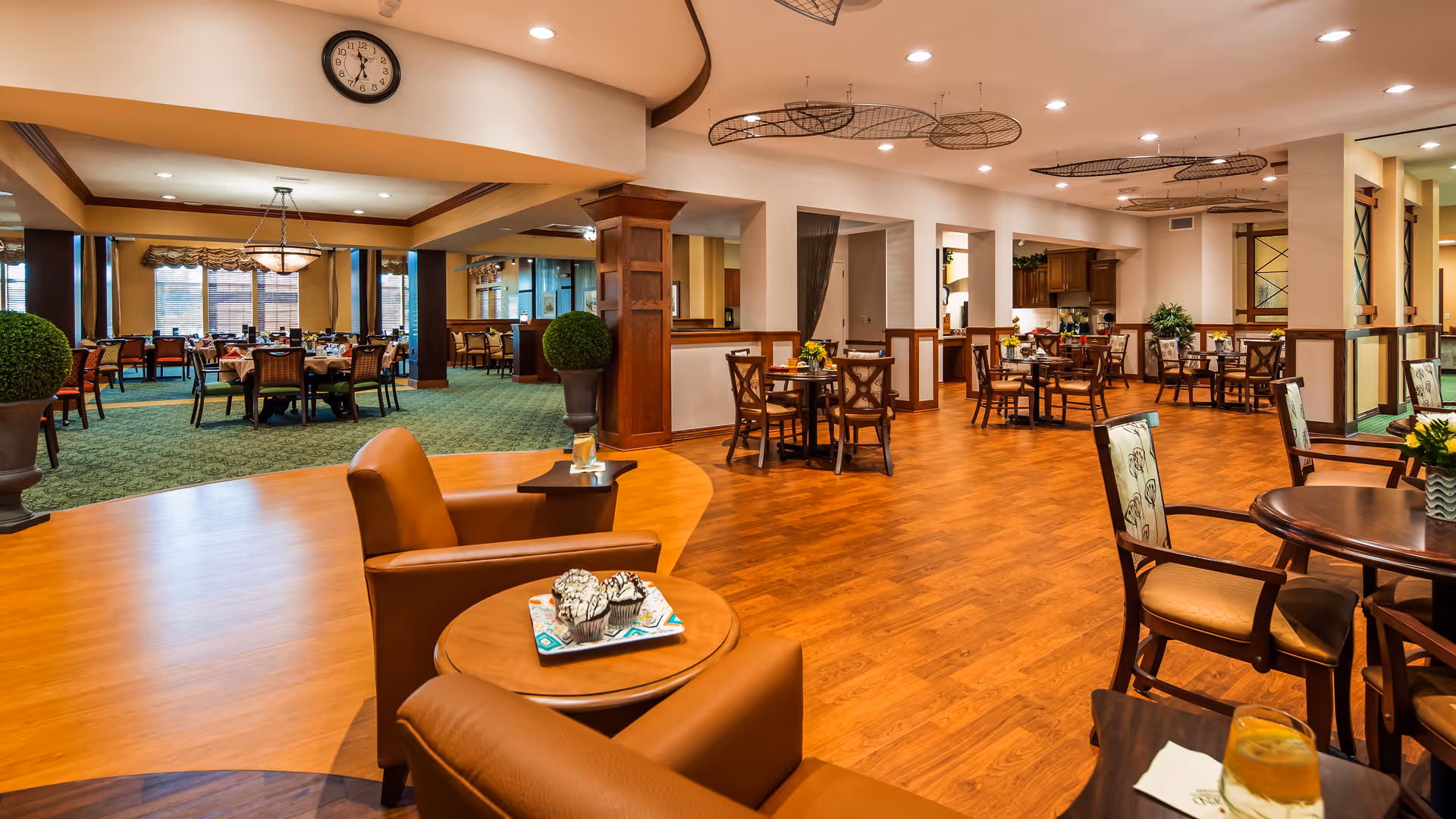 Interior view of a senior living facility dining and lounge area with wooden floors and carpeted sections. The space features multiple tables and chairs arranged for dining, comfortable armchairs with side tables, decorative plants, and warm lighting. A clock is mounted on the wall above the dining area.