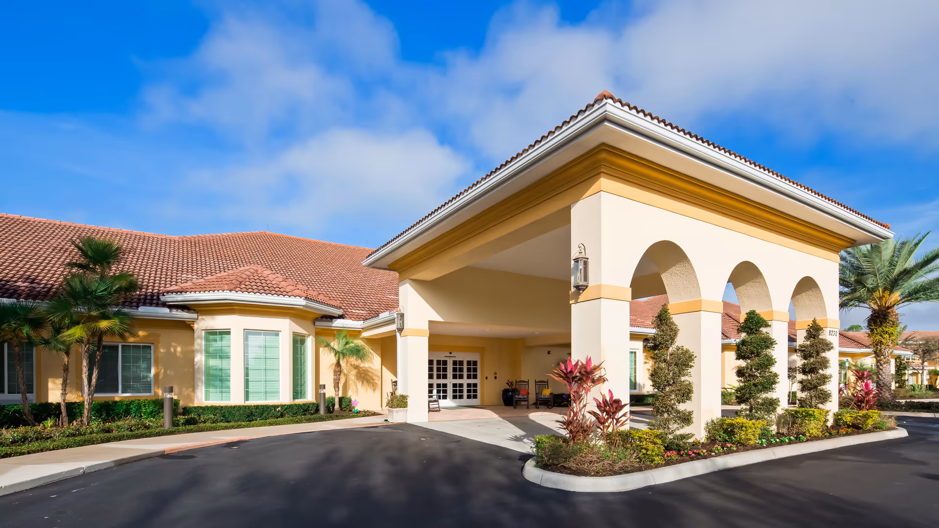 Exterior view of Windsor Reflections Memory Care Of Lakewood Ranch showing a building with a covered entrance supported by arches, surrounded by landscaped plants and palm trees under a blue sky with some clouds.